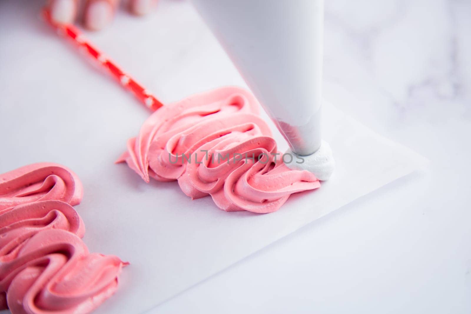 On light parchment there are pink meringue caps on sticks, the cook makes a pompom and a frill from a pastry bag of white meringue. Photography in a light key