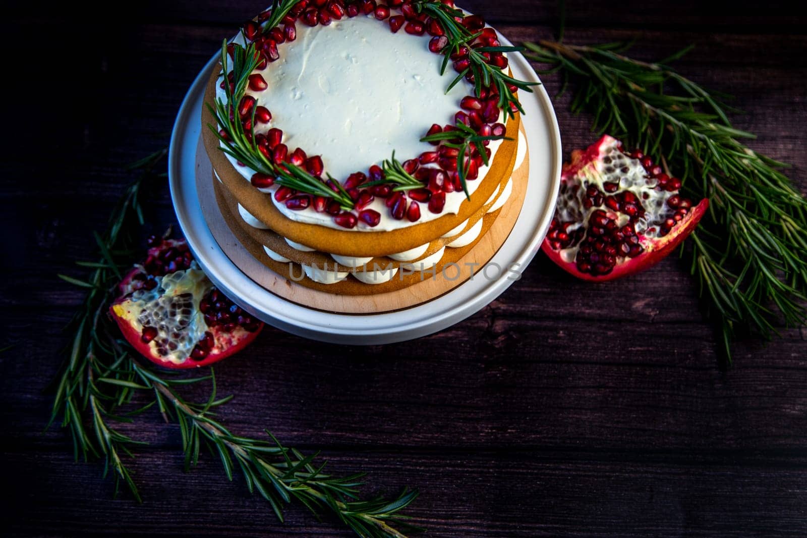 Cake with white cream and honey brown cakes lies on a white stand, decorated with pomegranate and rosemary, against a dark tree. High quality photo