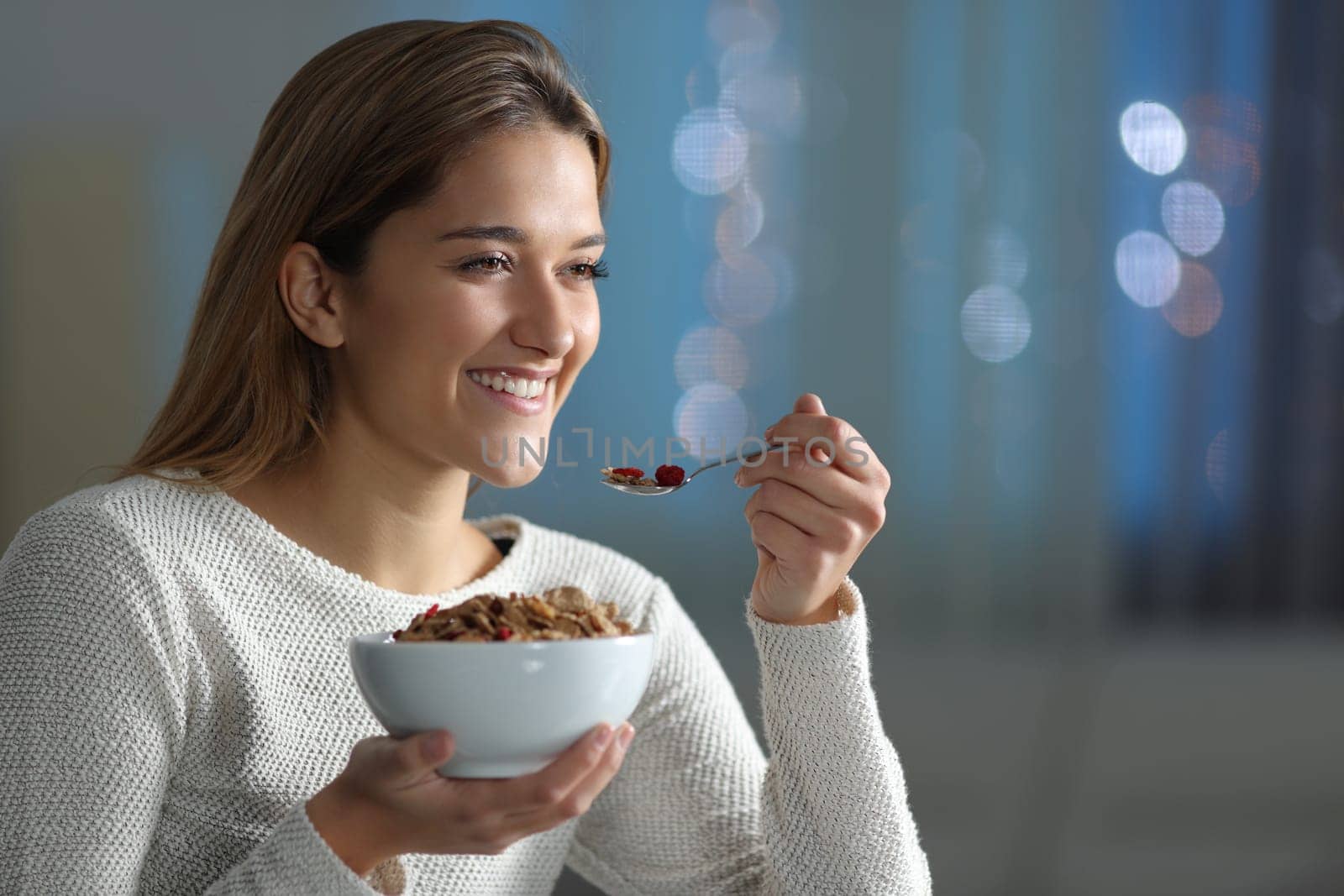 Happy woman eating cereal looking away in the night at home