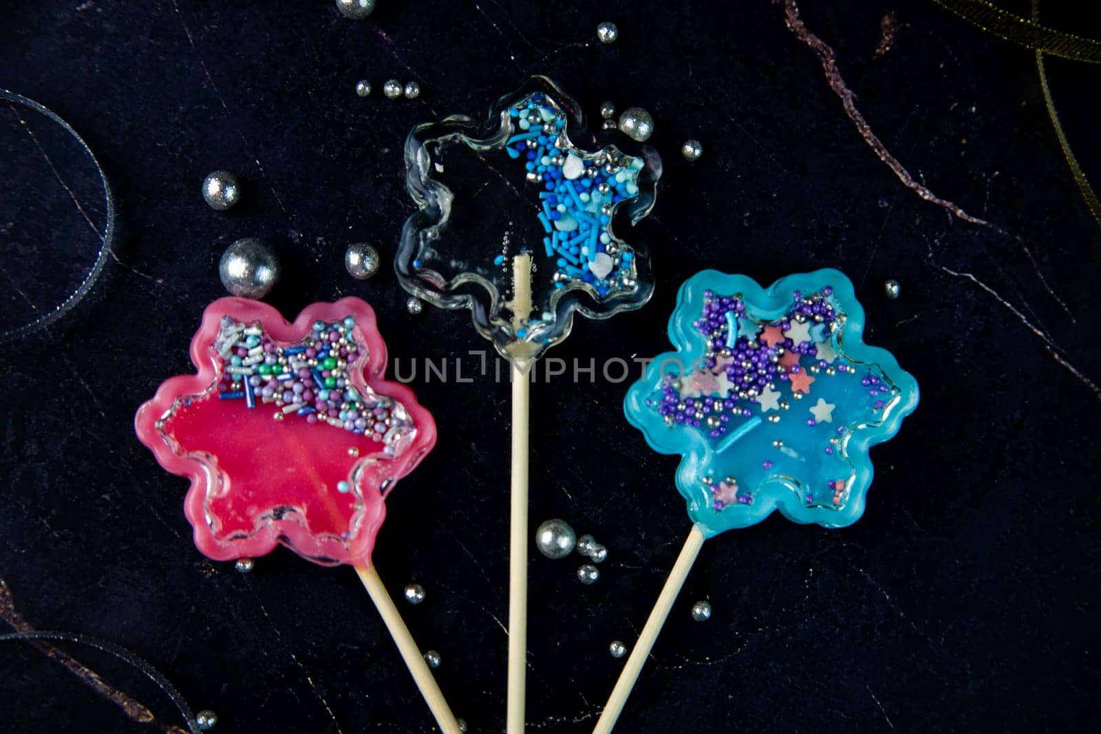 Lollipops on a wooden stick in the form of a rattle in the form of transparent, pink and blue snowflakes lie on a dark marble background surrounded by gold and silver ribbons and silver balls. High quality photo