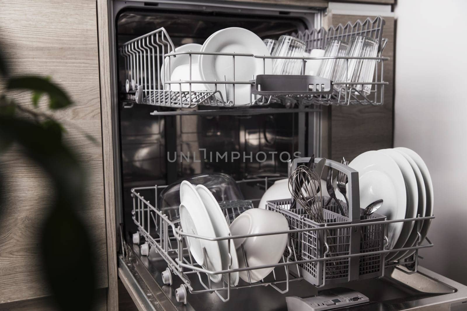 Front view of open automatic stainless steel built-in fully integrated top control dishwasher range machine with clean utensils, cutlery, glasses, dishes, plates inside in modern home kitchen
