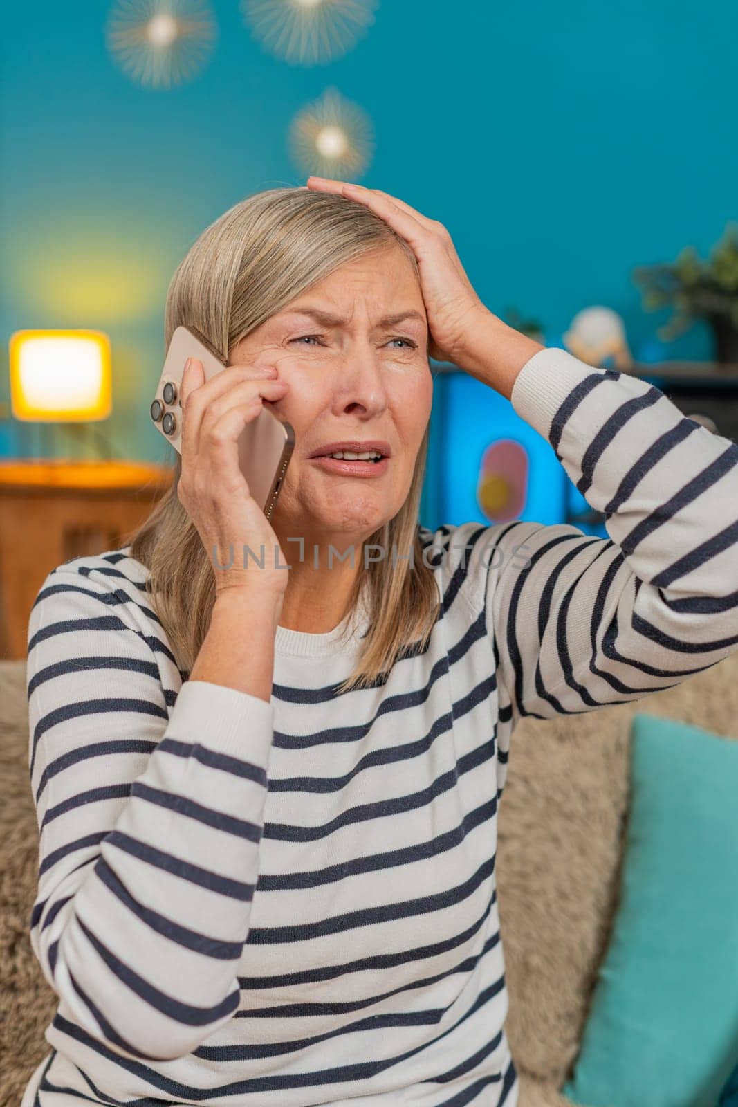 Mature upset anxious frustrated woman listening bad news talking on mobile phone. Grandmother sits on the sofa at home, holding the smartphone having conversation and showing signs of distress, stress