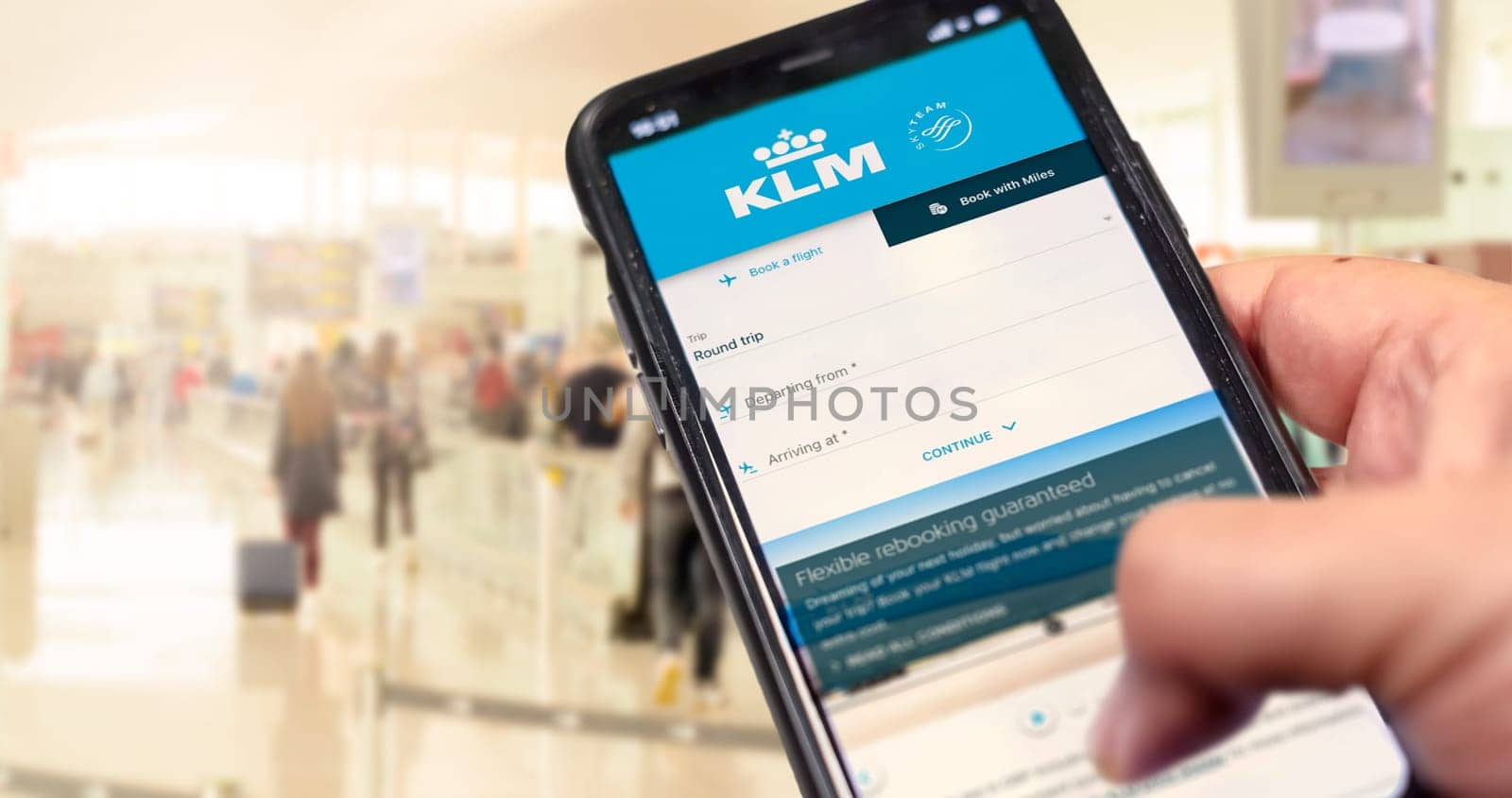 Amsterdam, NED, October 2021: Hand holding a phone with KLM Airlines flight booking application. Blurred airport with passengers in the background. Booking airline tickets for a round trip