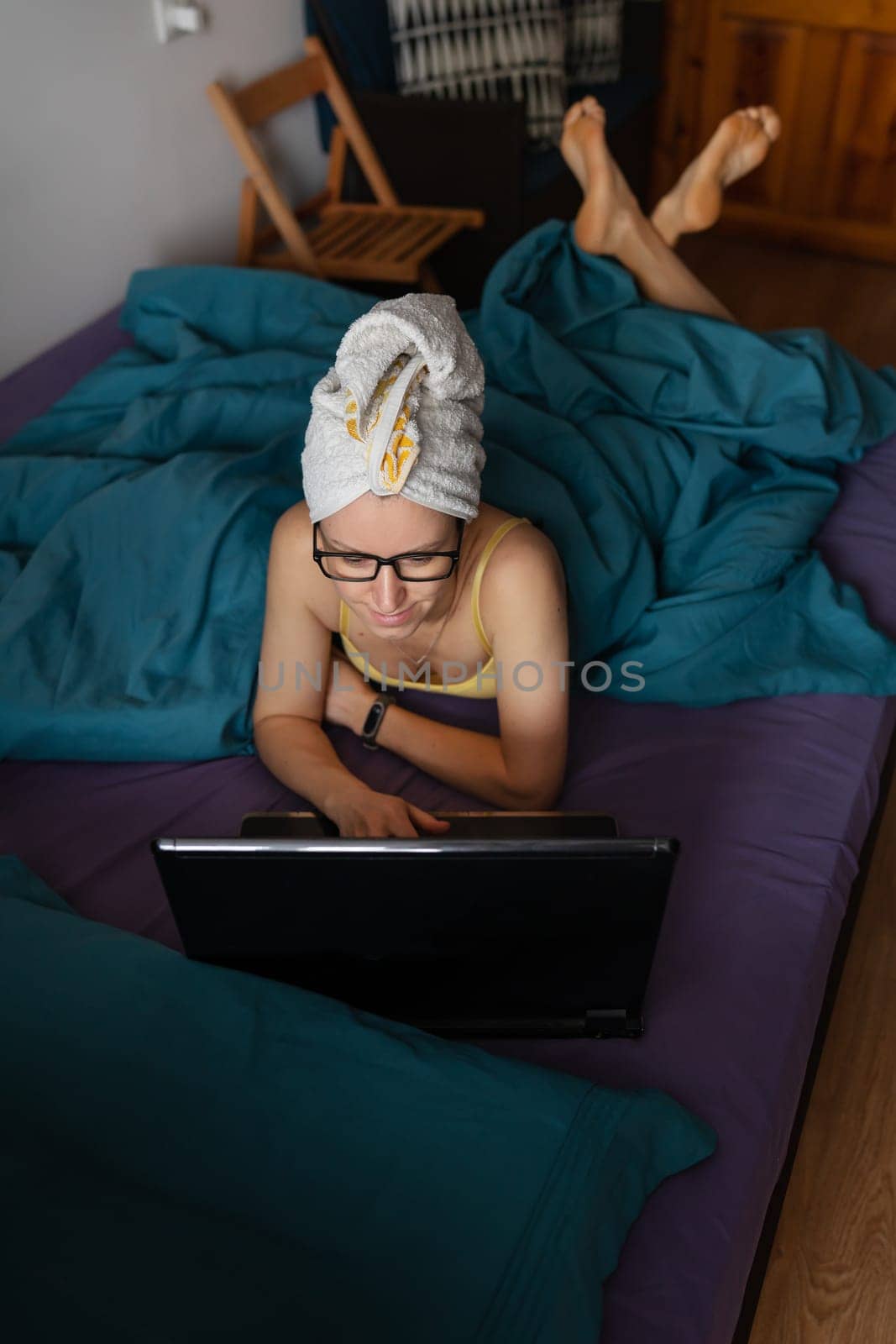 Home office, telecommuting, isolation, quarantine. A young girl working on a laptop lies under a blanket on a bed in a house in a towel after a shower