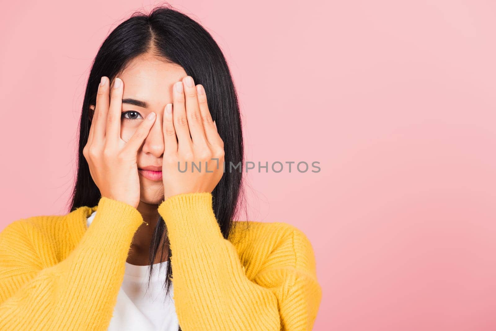Asian portrait beautiful young woman in depressed bad mood covering face with hands and peering out with one eye between her fingers studio shot isolated on pink background surprised and shocked