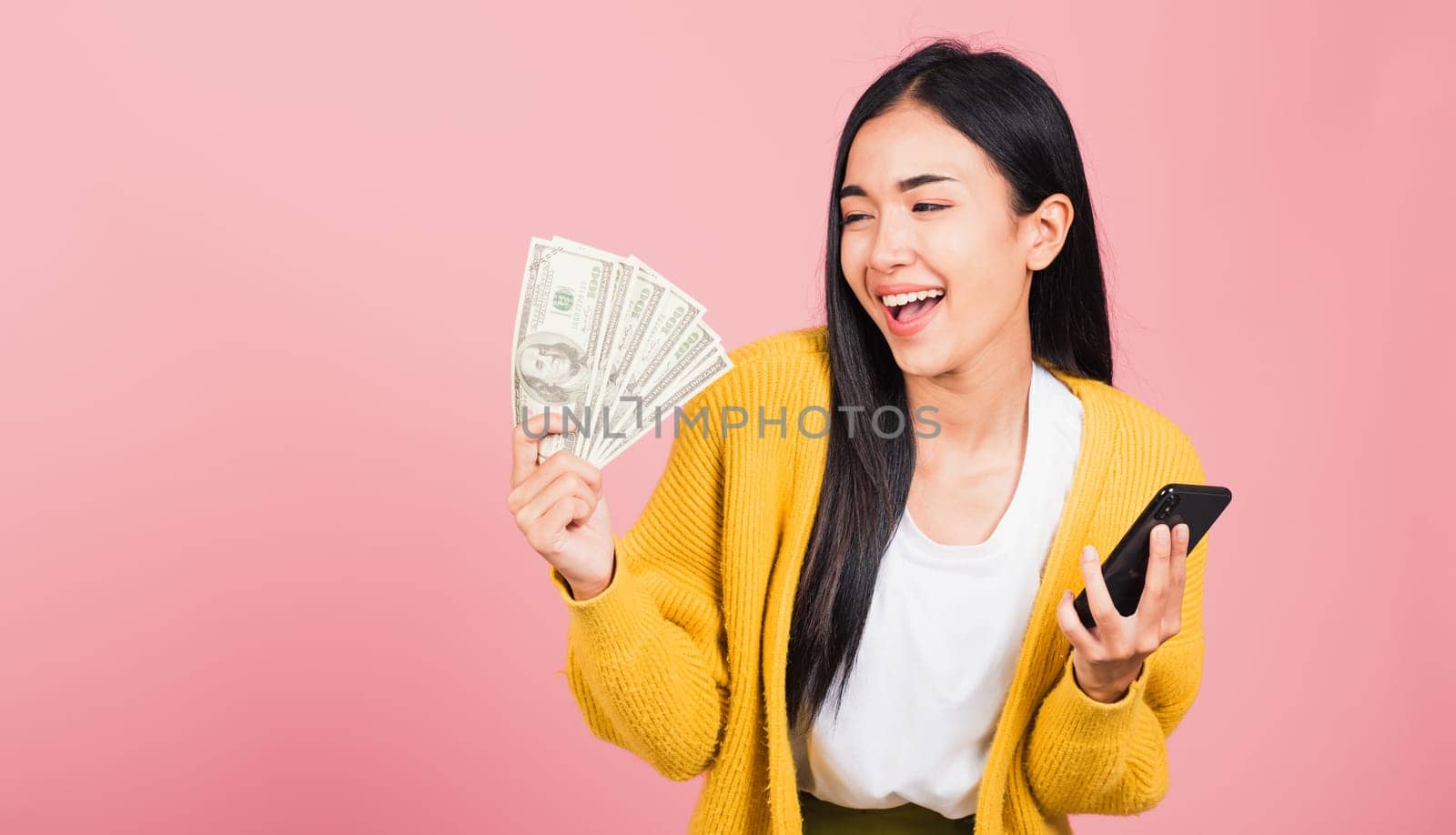 Portrait Asian happy beautiful young woman teen shopper smiling standing excited holding online smart mobile phone and dollar money banknotes on hand in summer, studio shot isolated on pink background