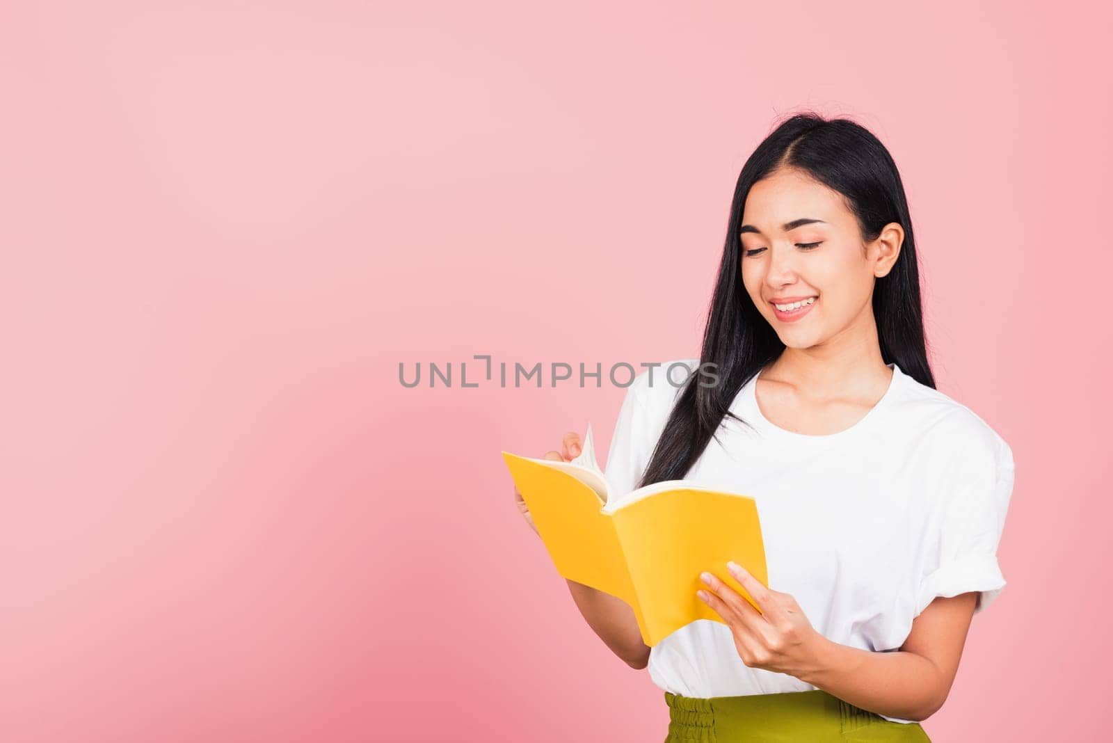 Portrait of beautiful Asian young woman teenage smiling holding a yellow book and reading, studio shot isolated on pink background with copy space