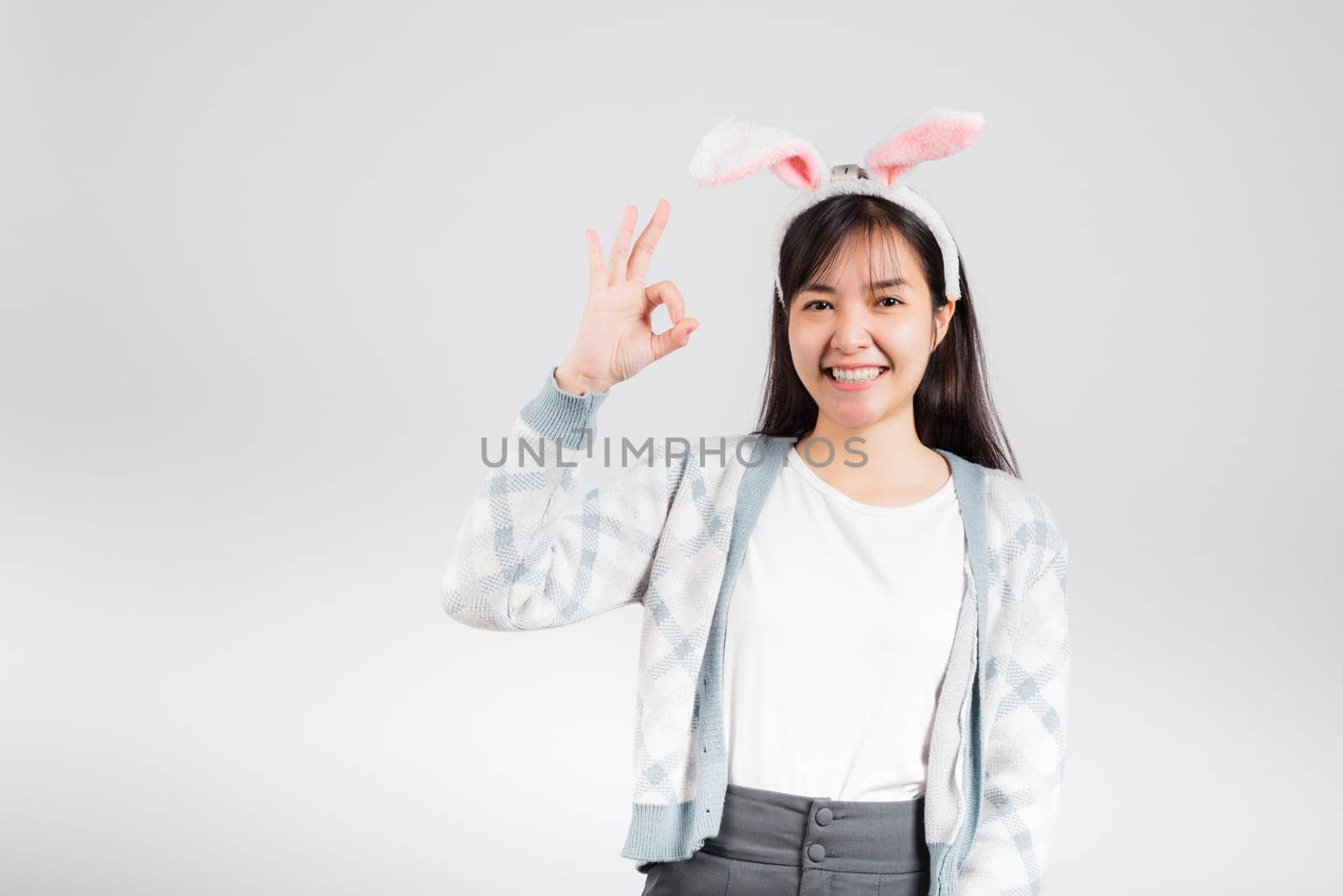 Easter day concept. Smiling happy woman wearing rabbit ears show OK finger sign, studio shot isolated on white background with copy space