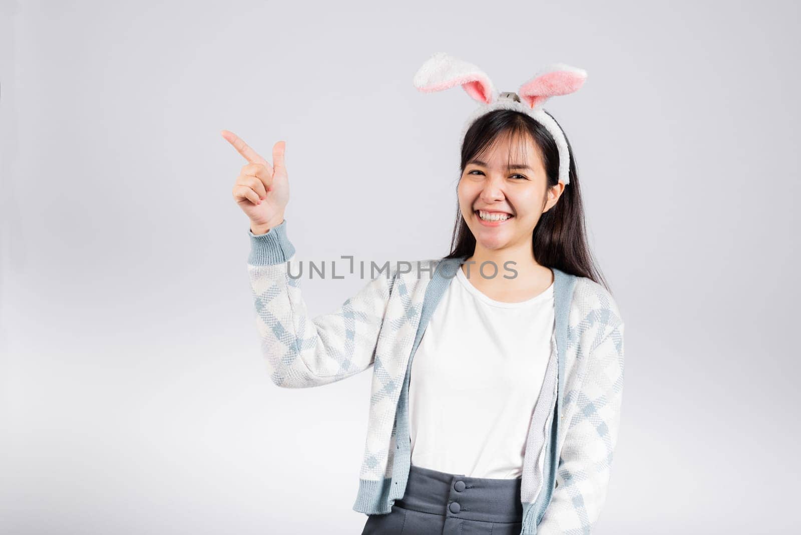 Easter day concept. Smiling happy woman wearing rabbit ears pointing finger something out space away side, studio shot isolated on white background with copy space