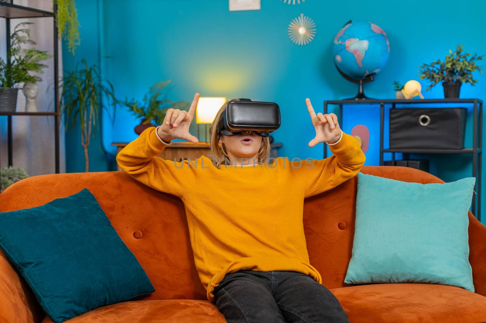 Caucasian teenager boy plays virtual reality game at home couch living room. Enthusiastically waves hands shows WOW emotion while sitting on sofa displaying excitement and enjoyment with VR technology