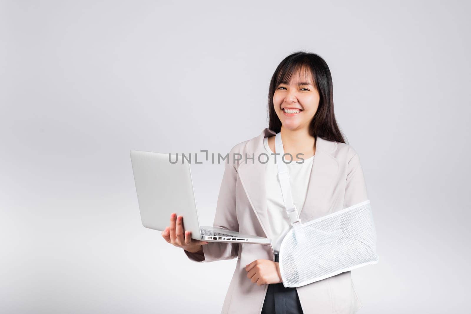 Asian business woman confident smiling broken arm after accident and wear arm splint for treatment but still have to work she hold laptop computer studio isolated on white background,copy space