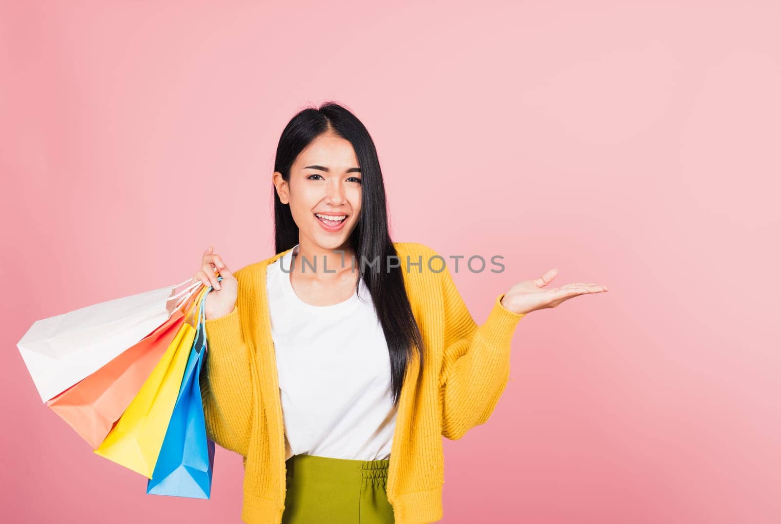 Portrait Asian happy beautiful young woman shopper smiling standing excited holding shopping bags multicolor in summer show something hand palm to copy space, studio shot isolated on pink background