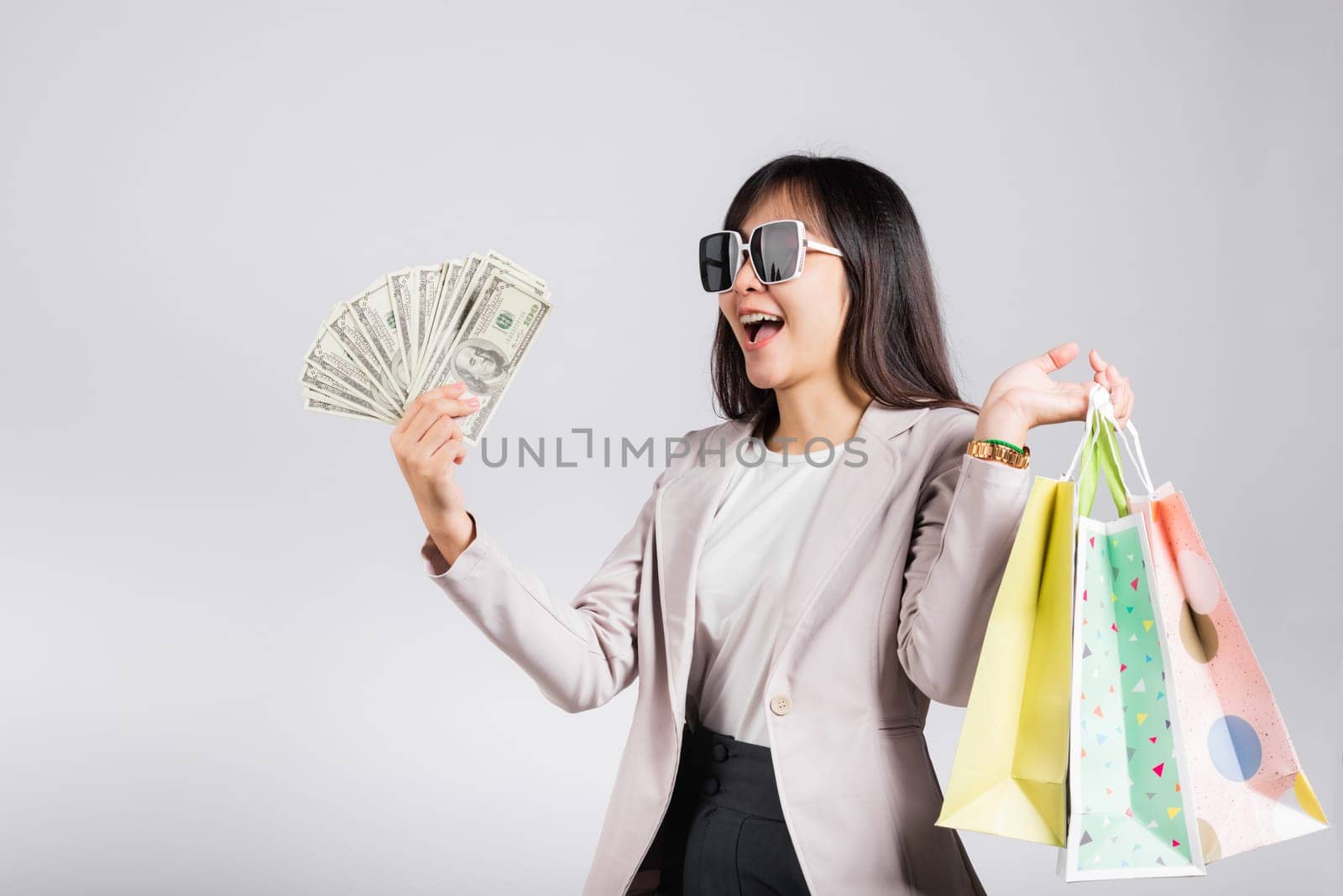 Woman with glasses confident shopper smile holding online shopping bags multicolor and dollar money banknotes on hand, excited happy Asian young female person studio shot isolated on white background