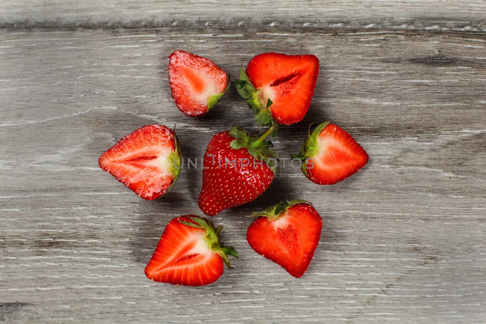 Tabletop view - whole strawberry with halves around, forming circle, on gray wood desk.