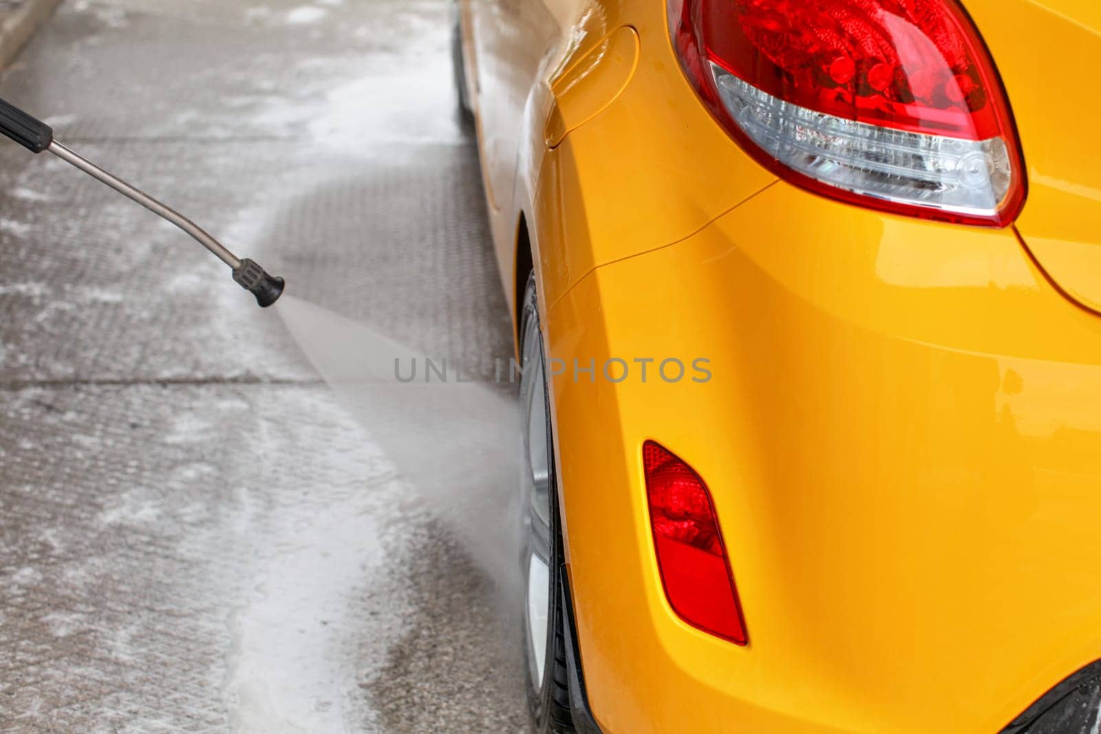 Rear wheel of yellow car washed with jet water in self serve carwash. View from behind.
