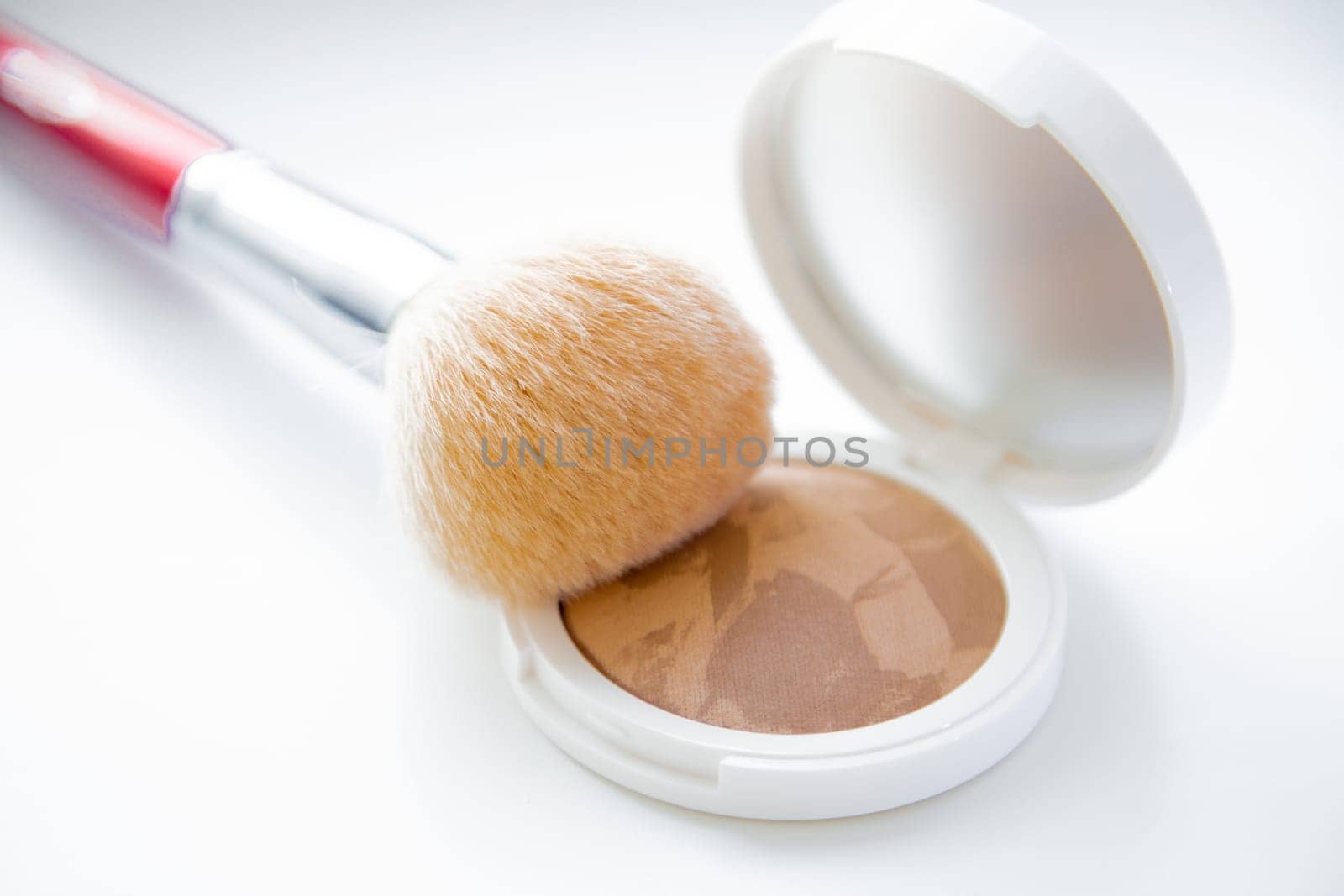 A white pillbox with powder and bronzer lies on a white background, on it lies a brush for blush on top. High quality photo