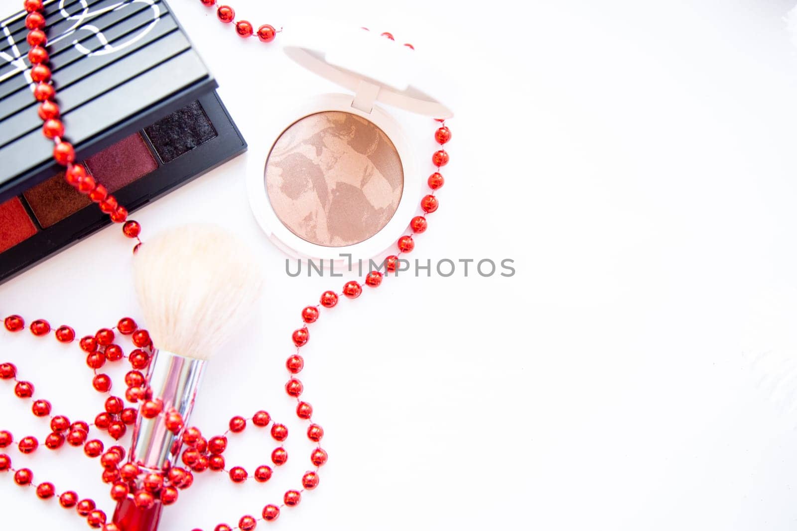 A white pillbox with powder and bronzer, a black package with eye shadow and a brush lie on a white background, they are entwined with red beads, with a place for text. High quality photo
