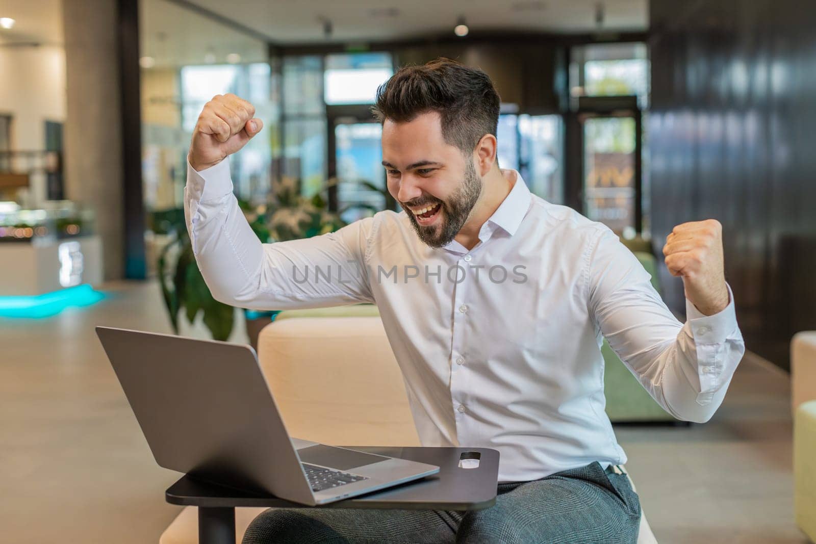 Caucasian bearded businessman working on laptop shocked by sudden victory, game winning lottery goal achievement, good career news. Excited guy freelancer clenching fists sitting in modern hotel lobby