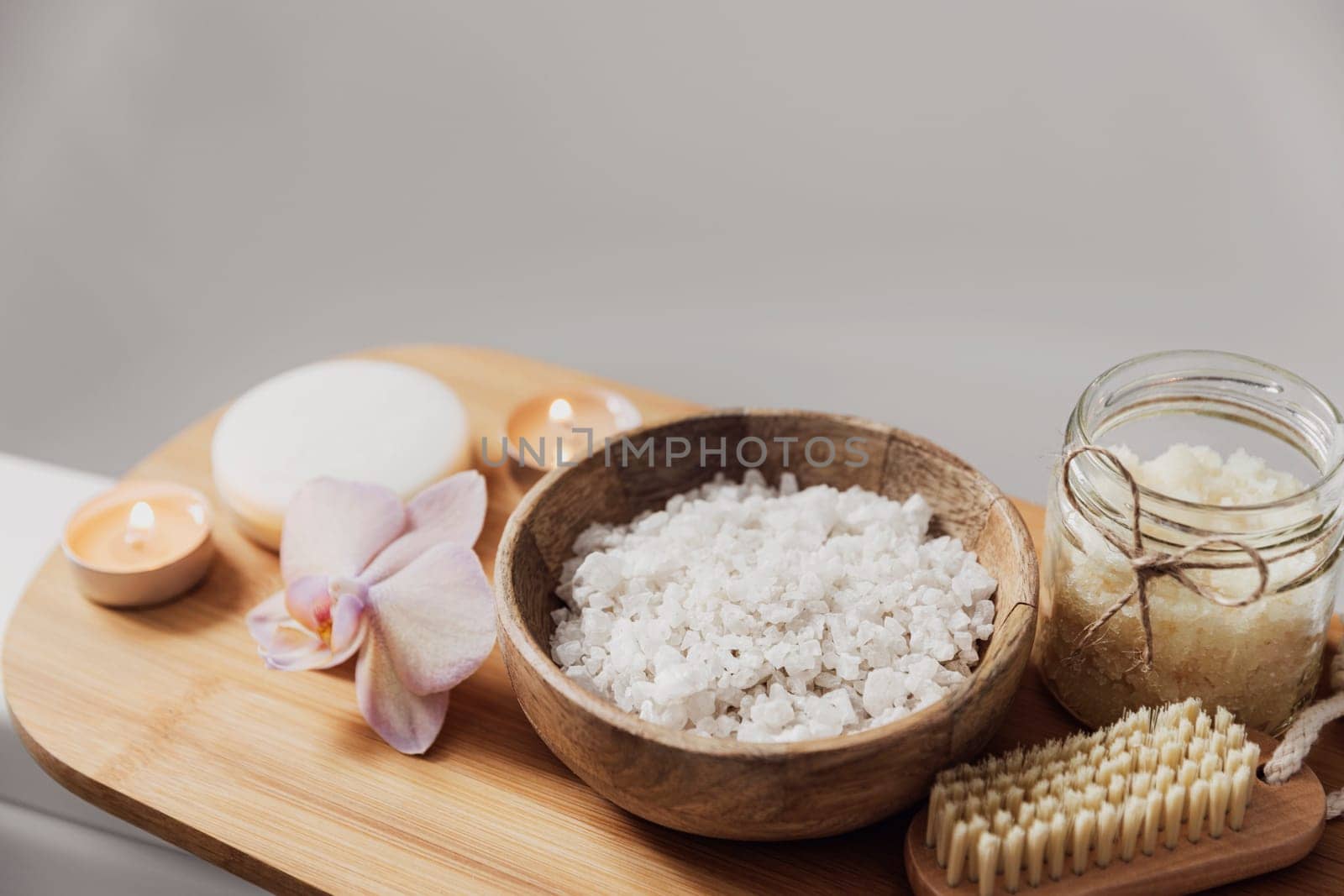 Preparation for hotel spa treatment or home bath procedure. White washbasin in bathroom with accessories on tray. Burning candles, washcloth, soap, foot brush, bottle with sea salt, orchid flower