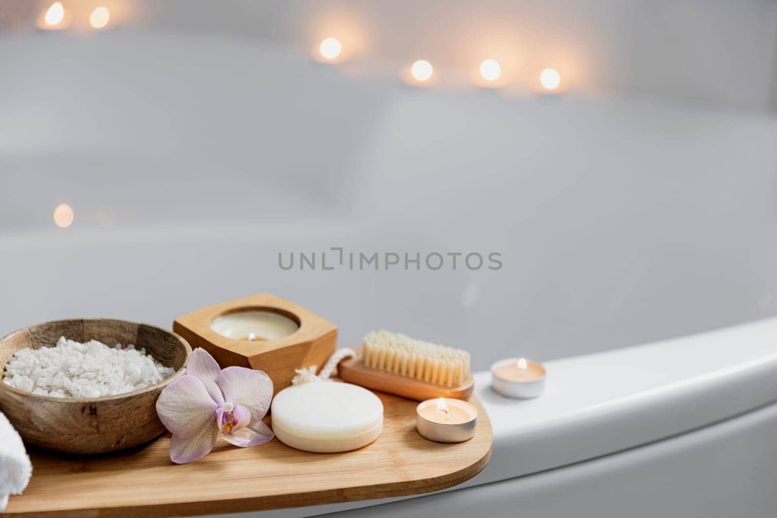 Preparation for hotel spa treatment, home bath procedure. White washbasin in bathroom, accessories on tray. Burning candles, soap, foot brush, towel, glass bottle with sea salt, orchid flower