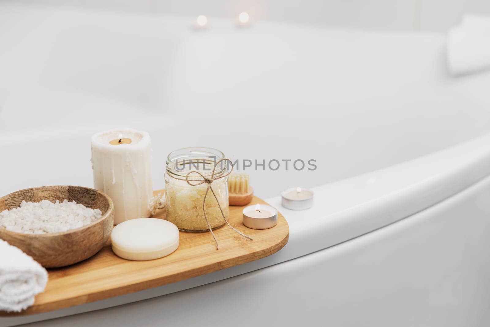 Preparation for hotel spa treatment, home bath procedure. White washbasin in bathroom, accessories on tray. Burning candles, soap, foot brush, towel, glass bottle with sea salt, orchid flower