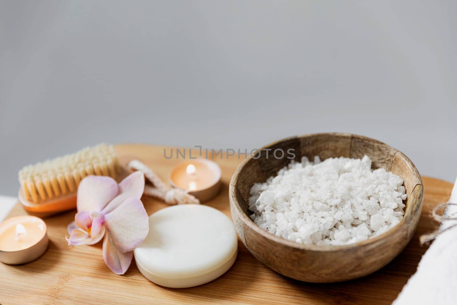 Preparation for hotel spa treatment or home bath procedure. White washbasin in bathroom with accessories on tray. Burning candles, washcloth, soap, foot brush, bottle with sea salt, orchid flower