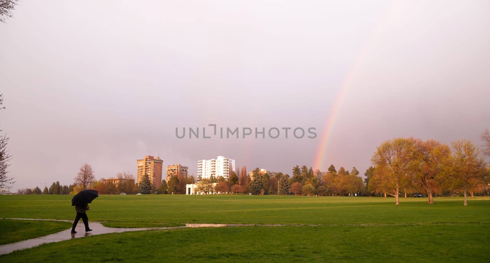 The sun peaks through for a moment lighting the way and producing a rainbow near a person walking with umbrella