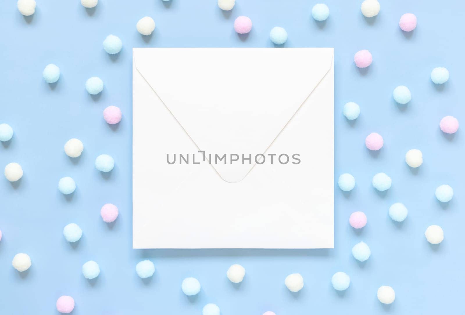 Blank envelope between pastel pom-poms on light blue top view. Romantic scene with blank horizontal card with place for text flat lay. Spring or boyish concept