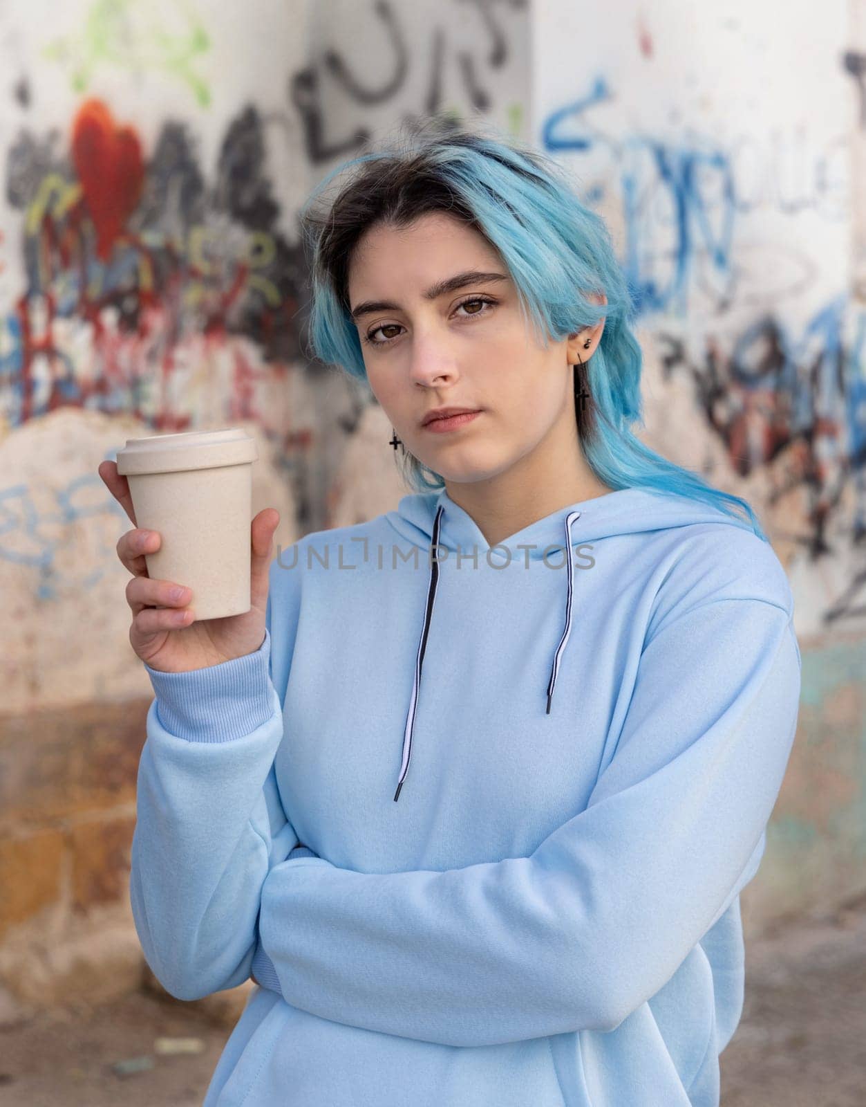 Teenage girl in light blue oversize hoodie with coffee to go and looking into the cameraBlue haired teen girl staying outdoors near graffiti wall. Clothing mockup. Hipster and adolescence concept