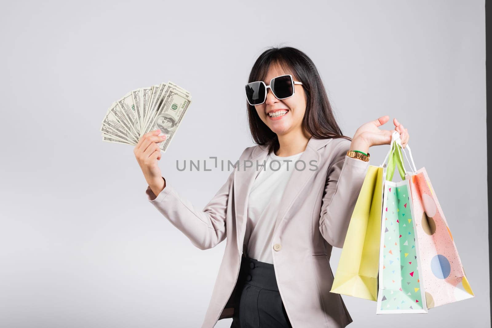 Woman with glasses confident shopper smile holding online shopping bags multicolor and dollar money banknotes on hand, excited happy Asian young female person studio shot isolated on white background
