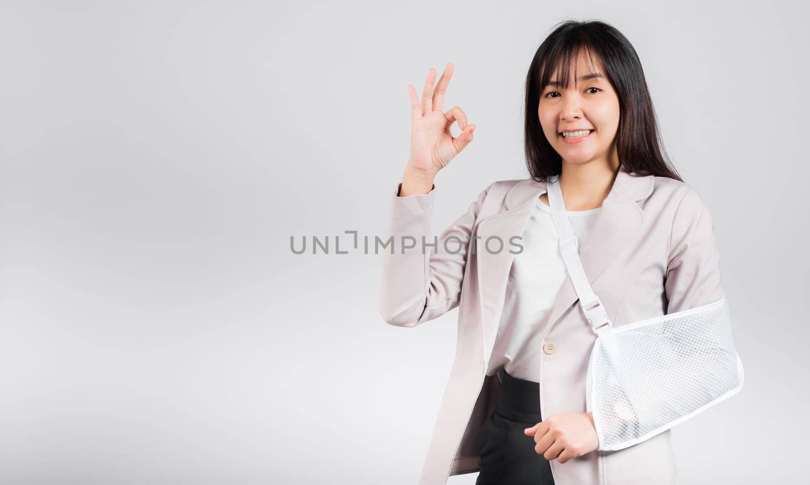 Arm broken. Business woman confident smile broken arm after accident wear arm splint for treatment show OK sign, happy Asian female sling support hand studio isolated on white background, copy space