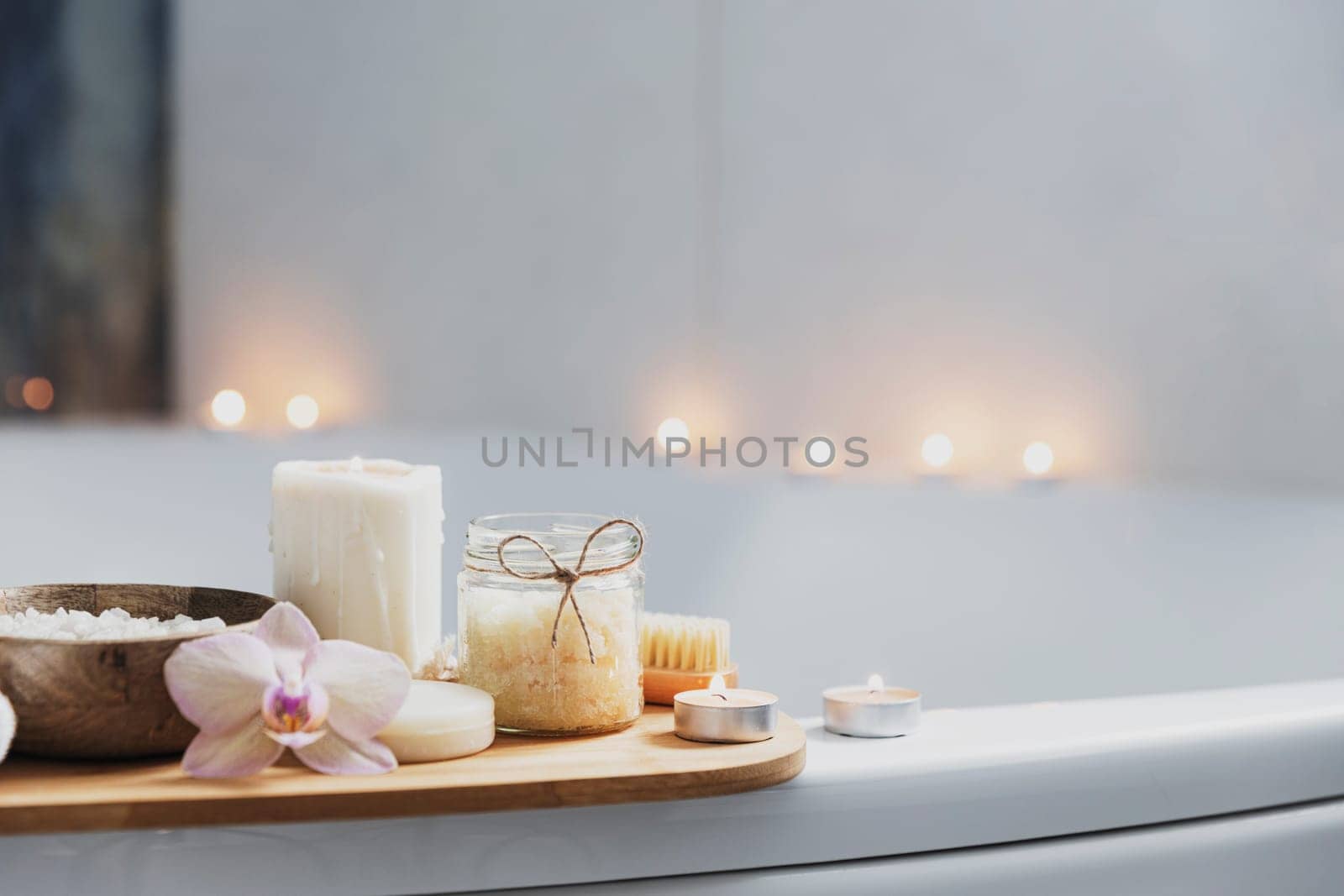 Preparation for hotel spa treatment or home bath procedure. White washbasin in bathroom with accessories on tray. Burning candles, washcloth, soap, foot brush, bottle with sea salt, orchid flower