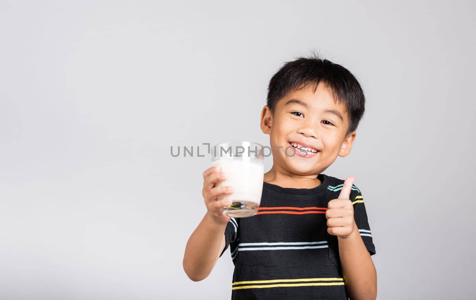 Little kid boy 5-6 years old smile holding milk glass he drinking white milk and show thumb up finger for good sign in studio shot isolated on white background, Daily life health care Medicine food