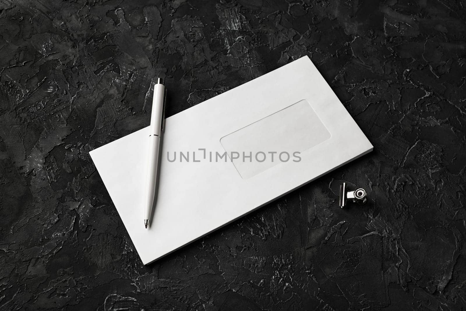 Photo of blank paper envelope, pen and clip on black stone background. Blank stationery template.