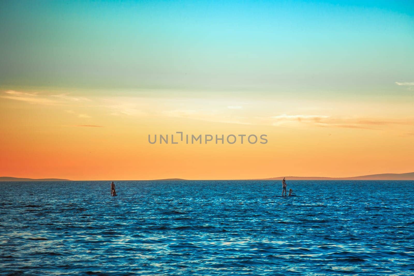 Sunset, blue sea and four people on a two sup boards on the seas. A beautiful sunset with blue and orange colors of the sky..
