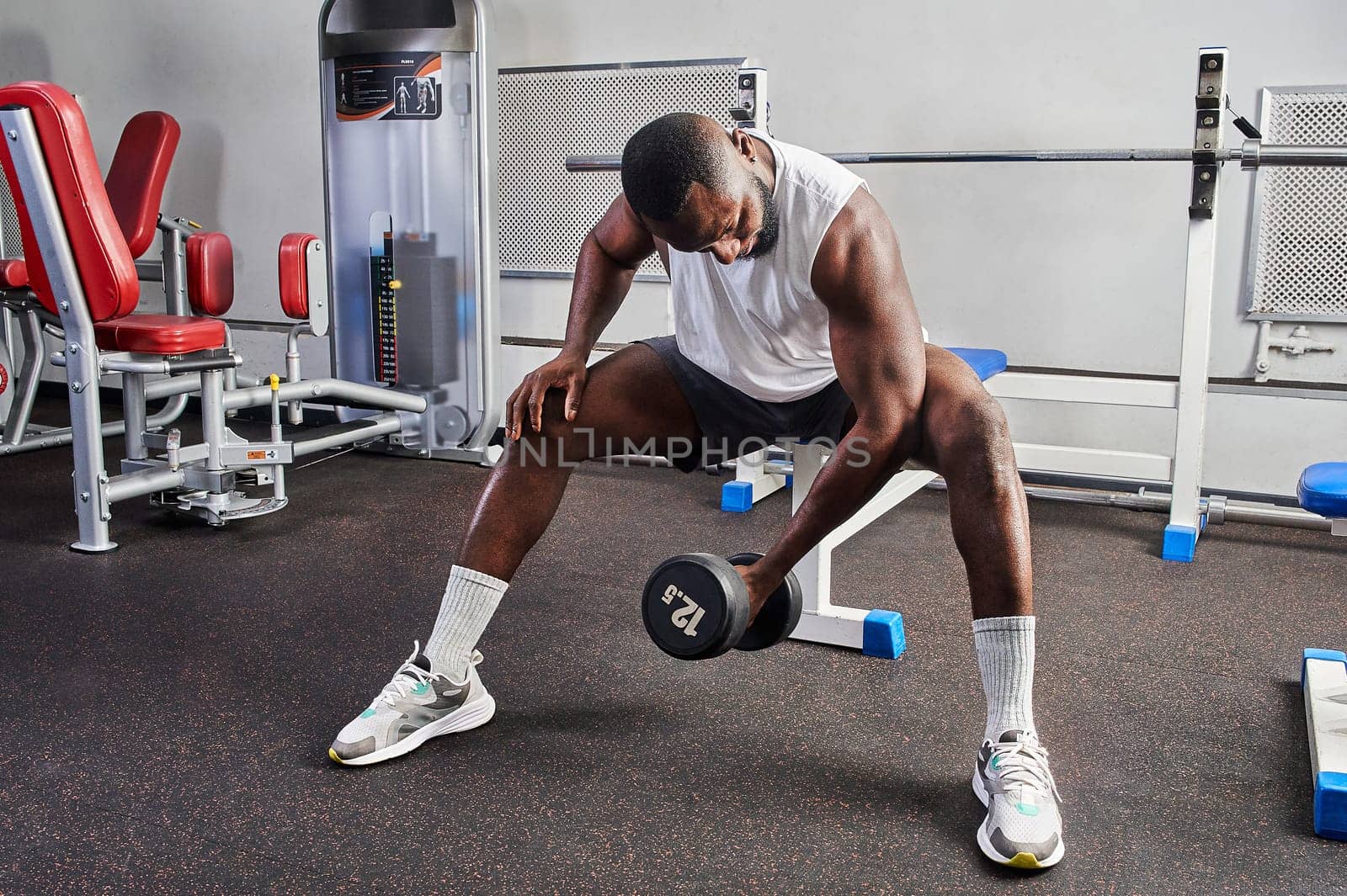 An African American man shakes his biceps while sitting on a bench in the gym. High quality photo