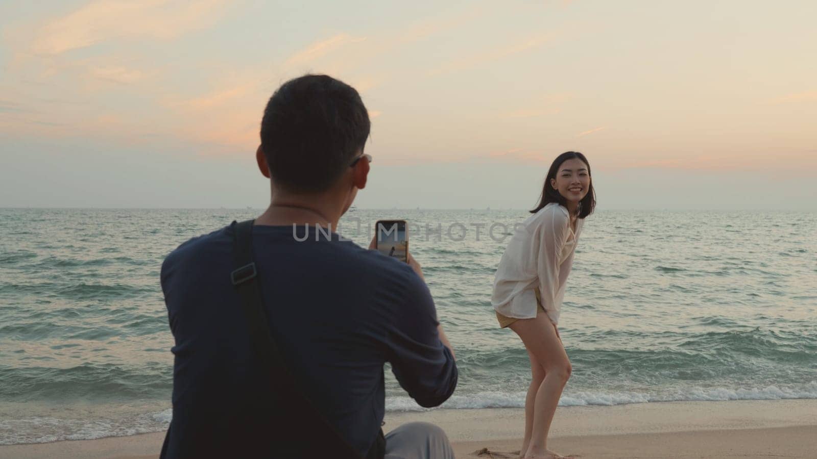 Happy man using smartphone camera making photo to woman at the beach and having fun together on beach in holiday vacation time at sunset, Young Asian woman and man in summer travel outdoor