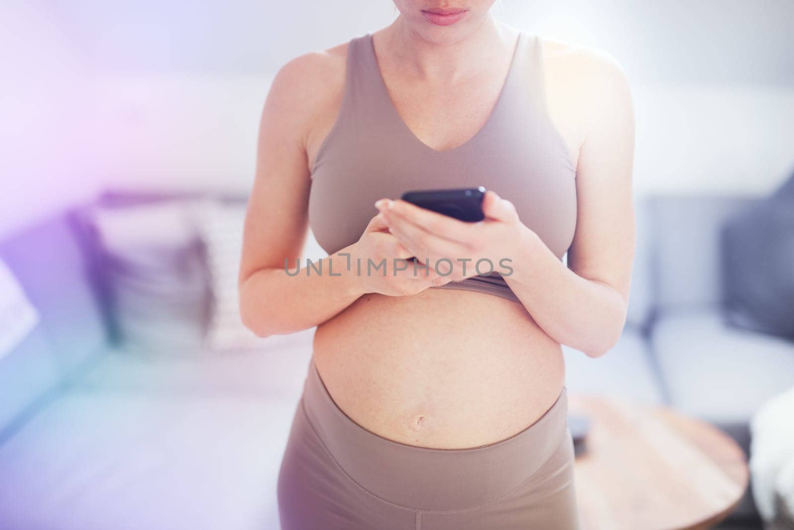 Closeup photo of pregnant female belly. Woman holding and using mobile smart phone app at home interiors. Pregnancy, technology, online shopping, preparation and expectation concept.