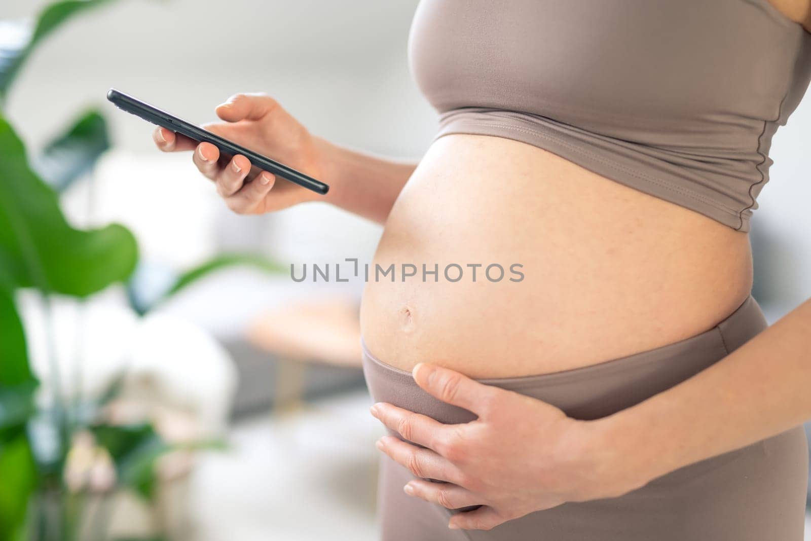 Closeup photo of pregnant female belly. Woman holding and using mobile smart phone app at home interiors. Pregnancy, technology, online shopping, preparation and expectation concept.