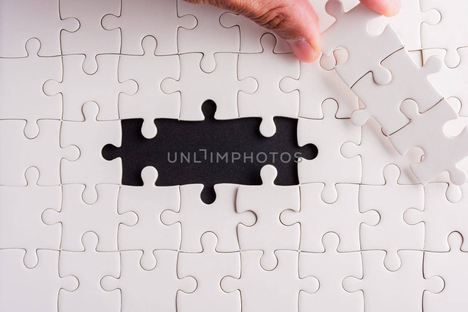 Top view flat lay of hand-holding last piece white paper jigsaw puzzle game last pieces put to place for solve problem complete mission, studio shot on a black background, quiz calculation concept