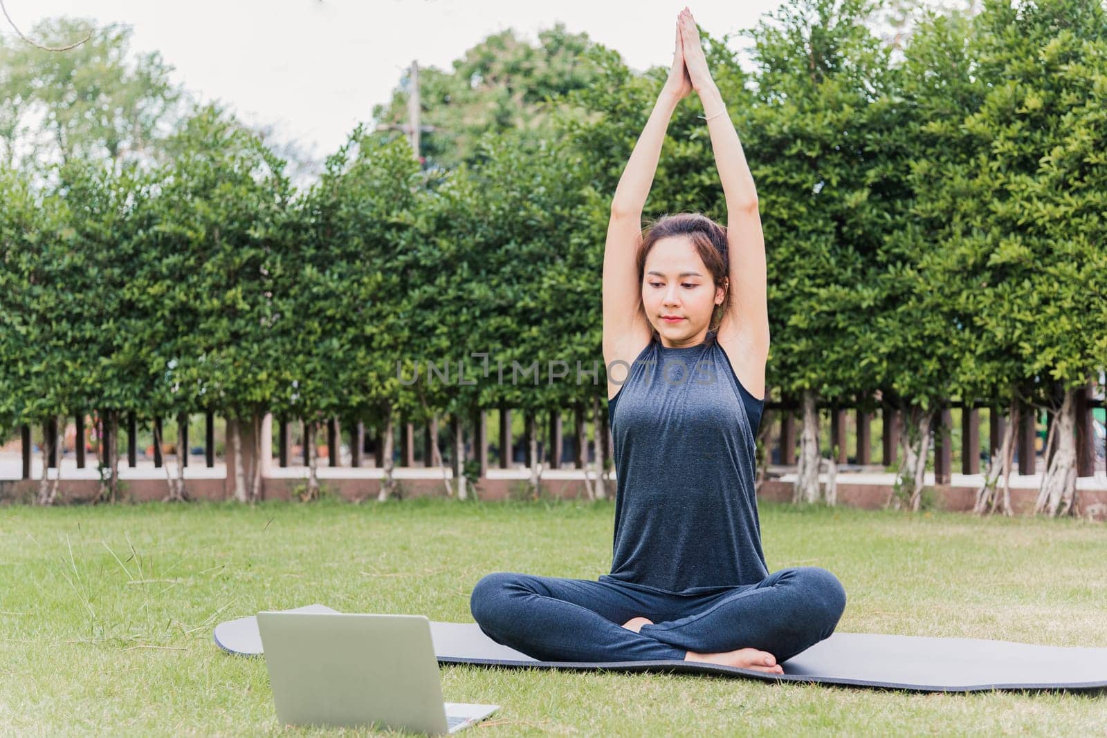 Asian young woman using laptop during online video training practicing yoga outdoors in meditate pose sitting on green grass in nature a field garden park, Stretching health care concept
