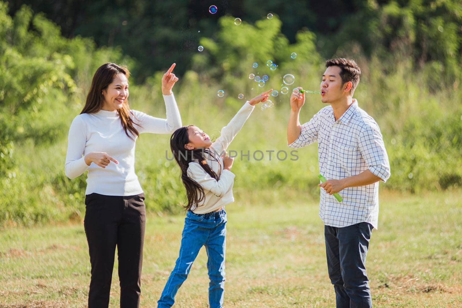Happy Asian lifestyle family mother, father and little cute girl child having fun together and enjoying outdoor play blowing soap bubbles in the garden park on a sunny day, summertime