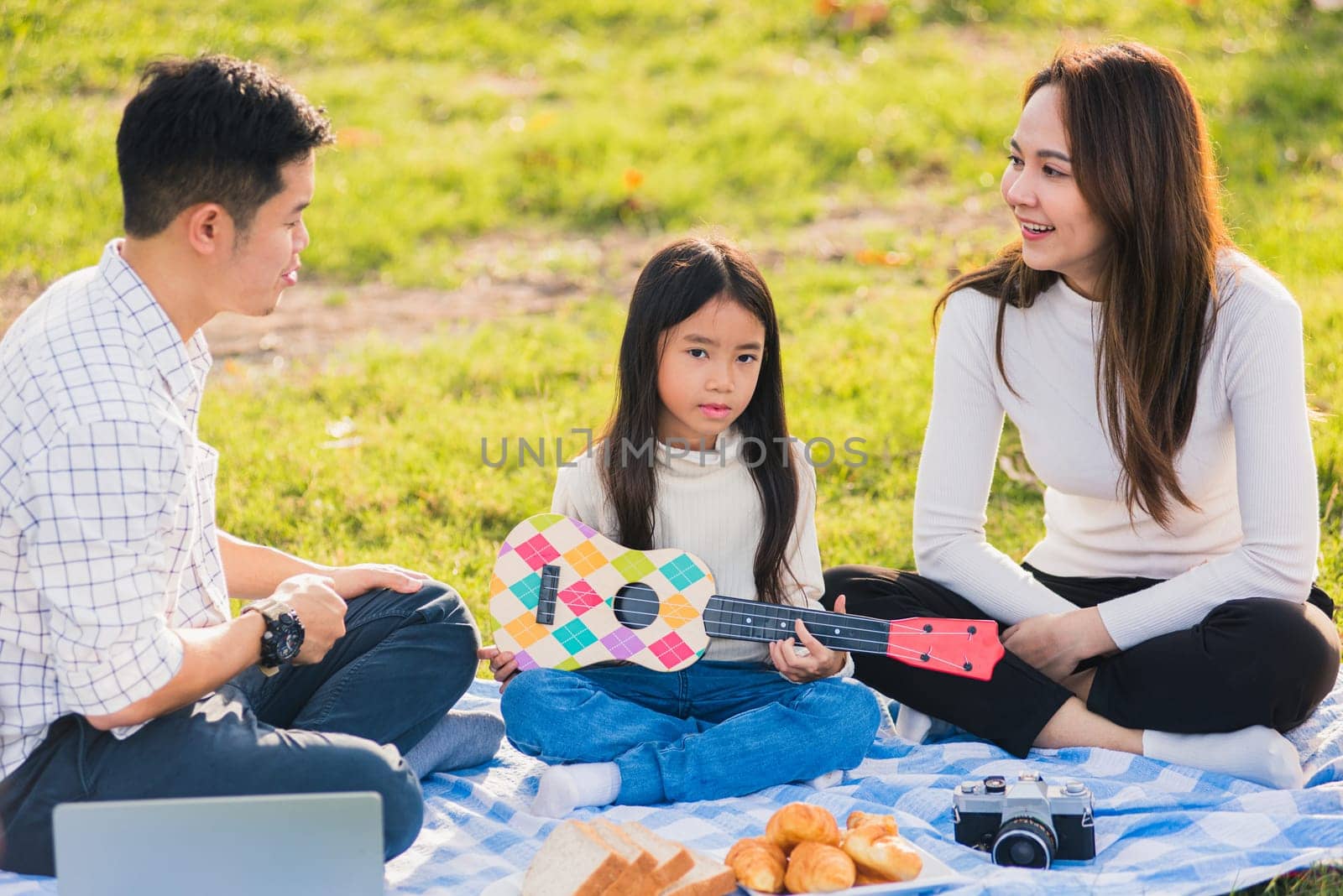 Happy Asian young family father, mother and children having fun and enjoying outdoor together sitting on the grass party with playing Ukulele during a picnic in the garden park on a sunny day