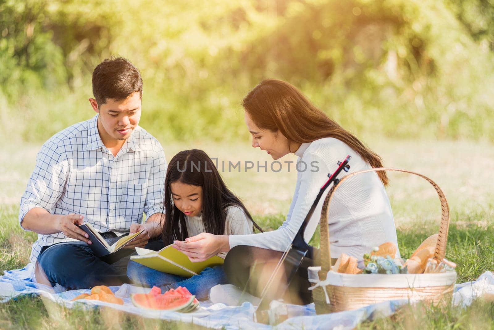 Happy Asian young family father, mother and child little girl having fun and enjoying outdoor on picnic blanket reading book in park at sunny time, summer leisure spring concept