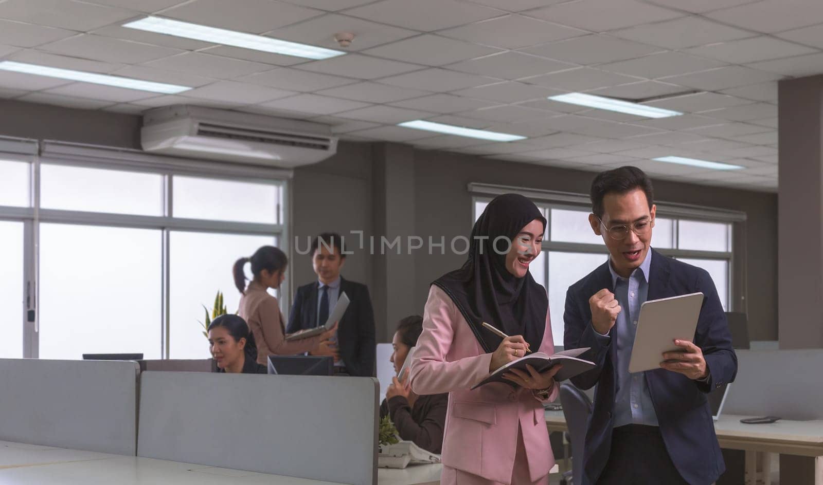 Happy businessman in dark blue suit using digital tablet working with Muslim women wearing hijab making notes while standing in open space office. Business team workplace concept
