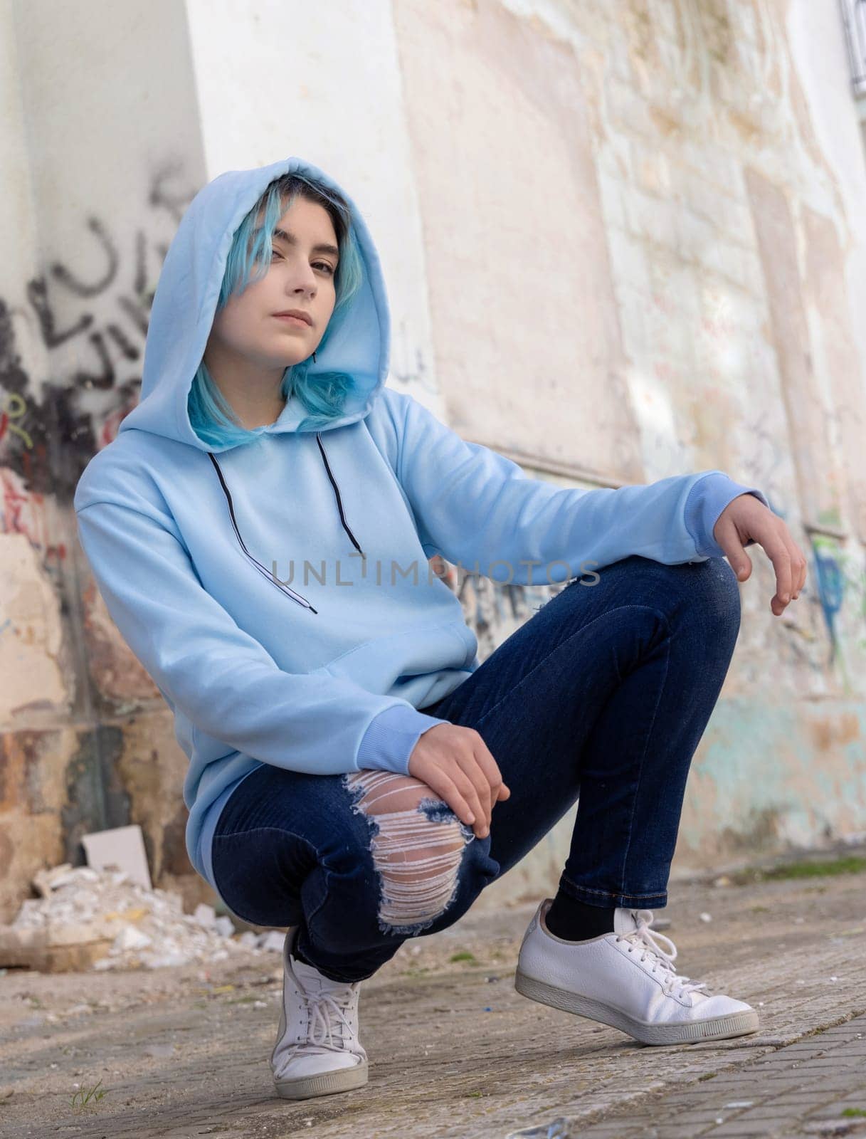 Blue haired Teenage girl in light blue oversize hoodie and looking aside. Blue haired teen girl sitting on her haunches near graffiti wall. Clothing mockup