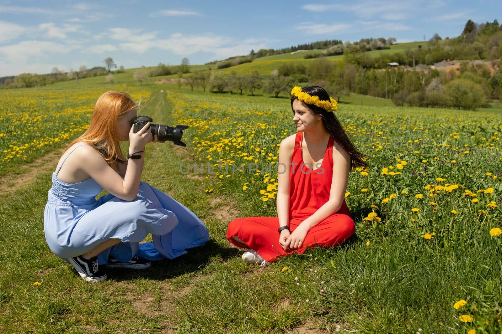Young girl posing photographing friend sitting in a dandelion meadow.