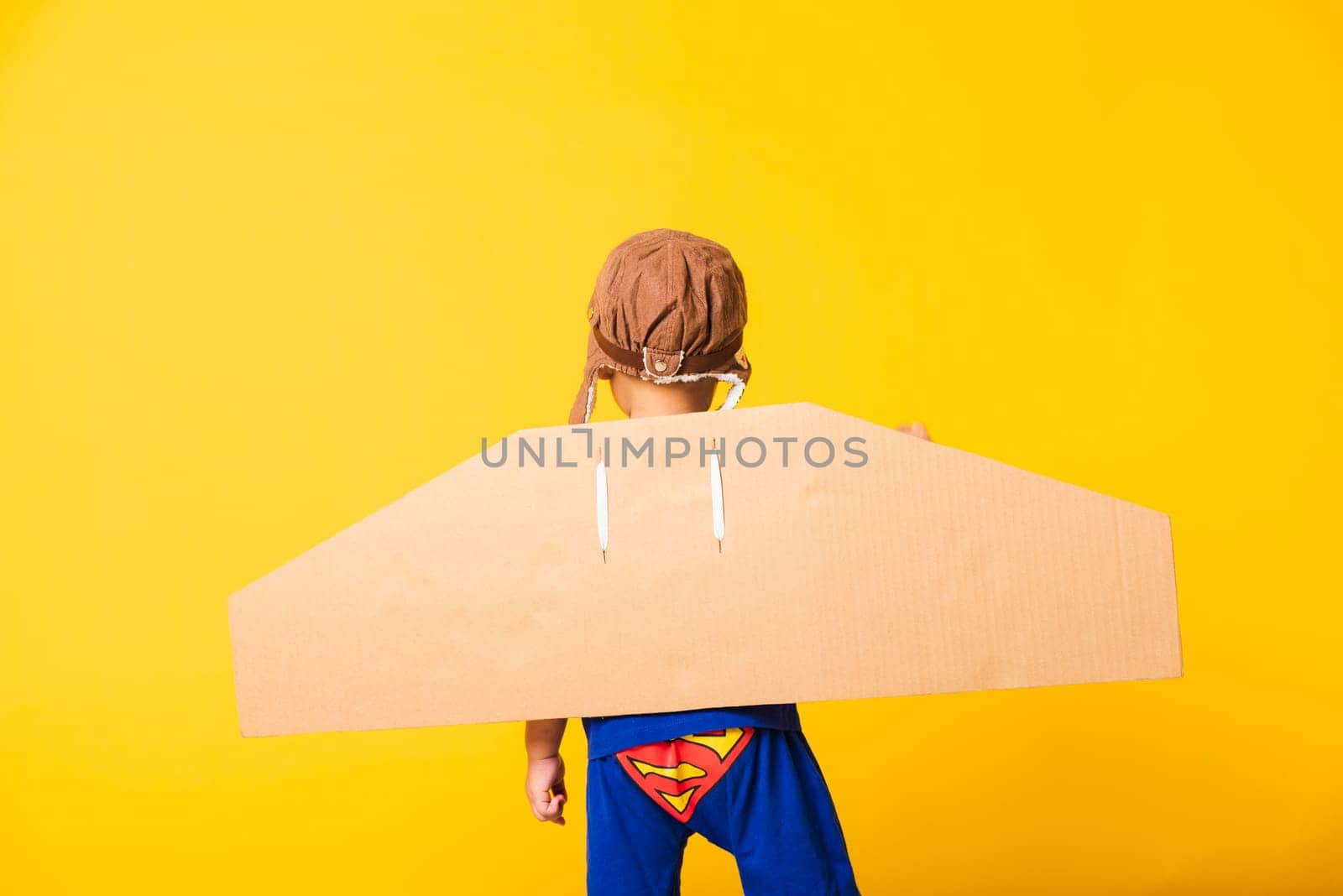 Back of Happy Asian handsome funny child or kid little boy smile wear pilot hat play raise hand up with toy cardboard airplane wings flying, studio shot isolated yellow background, Startup freedom