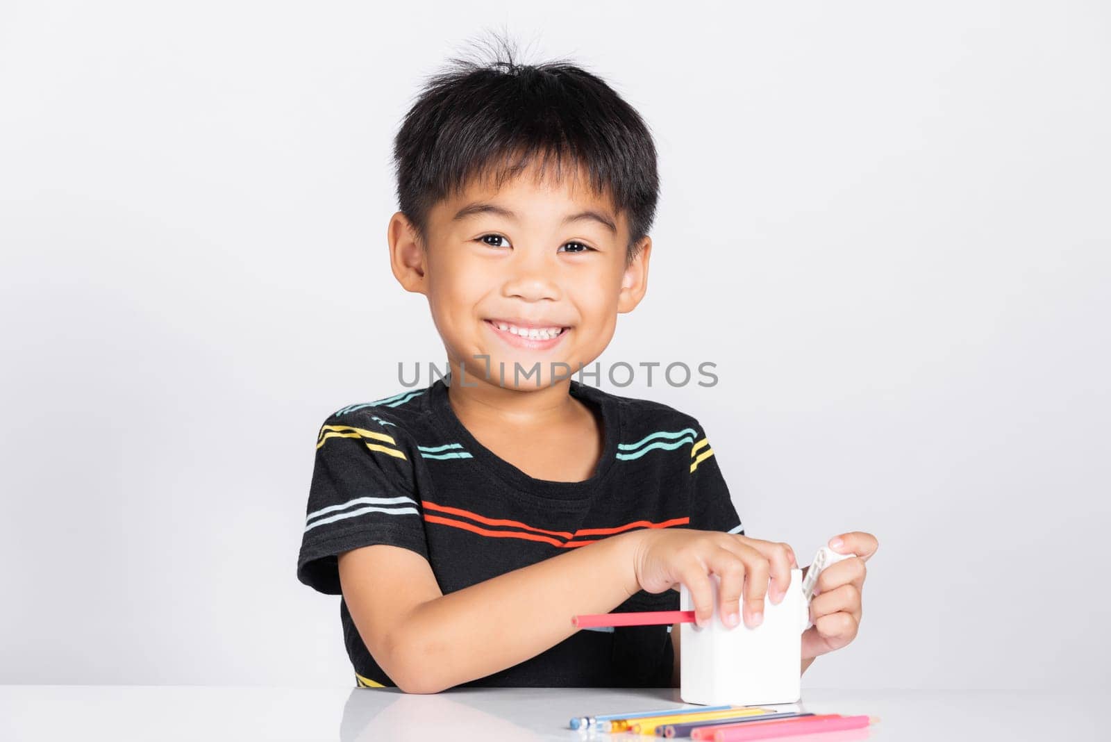 Little cute kid boy 5-6 years old smile using pencil sharpener while doing homework in studio shot isolated on white background, Asian children preschool sharpening color pencils, education concept