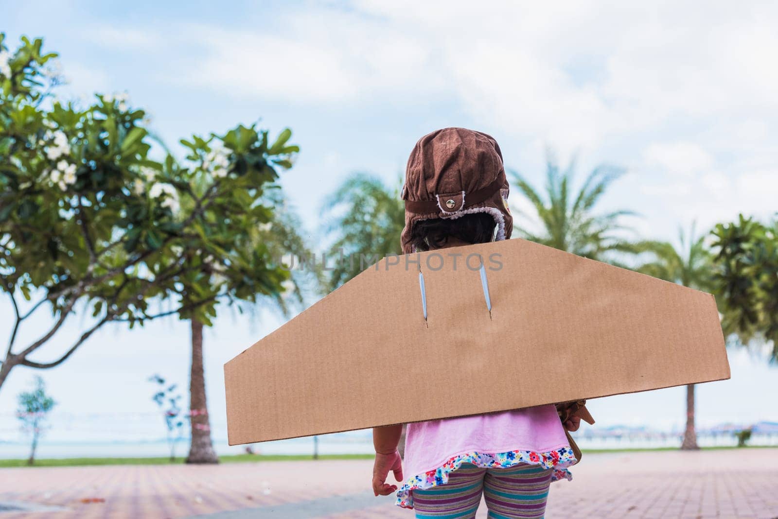 Happy Asian funny child or kid little girl smile wear pilot hat and goggles play toy cardboard airplane wing flying against summer sky cloud on trees garden background, Startup freedom concept